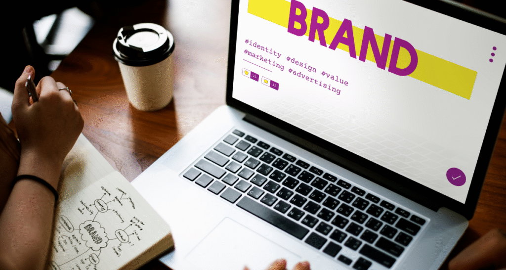 "Person working on a laptop with a coffee cup, viewing a screen with 'BRAND' and hashtags like #identity, #design, and #marketing, while brainstorming visual identity concepts in a notebook."